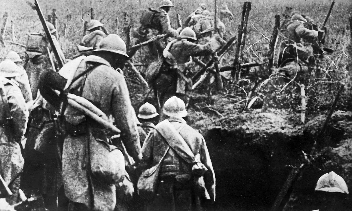 Black-and-white photo of French soldiers with packs and rifles climbing out of a trench into a field of shattered wire posts and smoke.