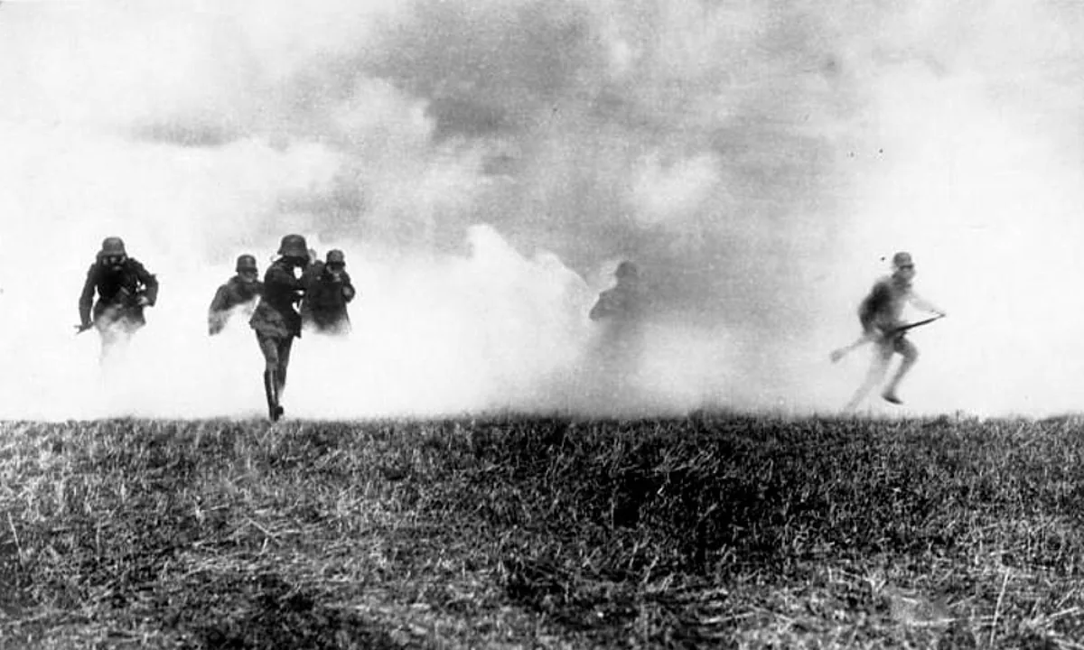 Black-and-white photo of helmeted soldiers wearing gas masks running out of a thick white cloud across a grassy field.
