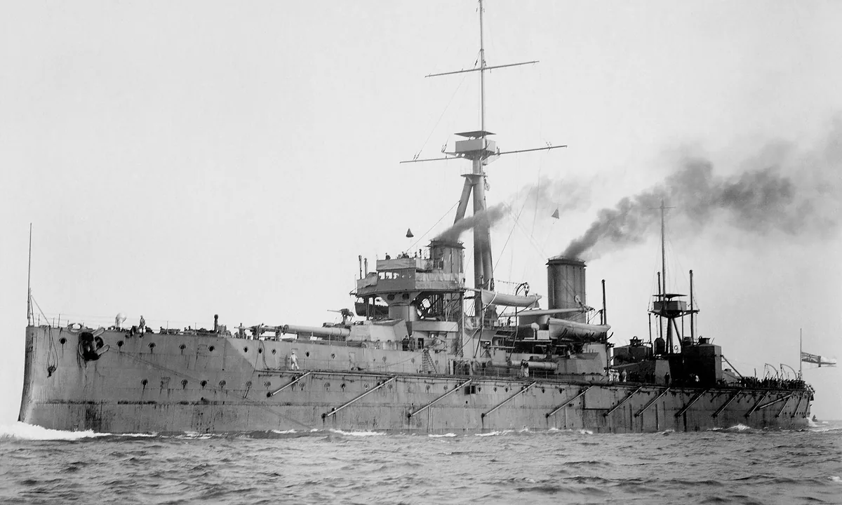 Black-and-white photo of a large grey battleship with tripod mast and two funnels steaming through choppy water.
