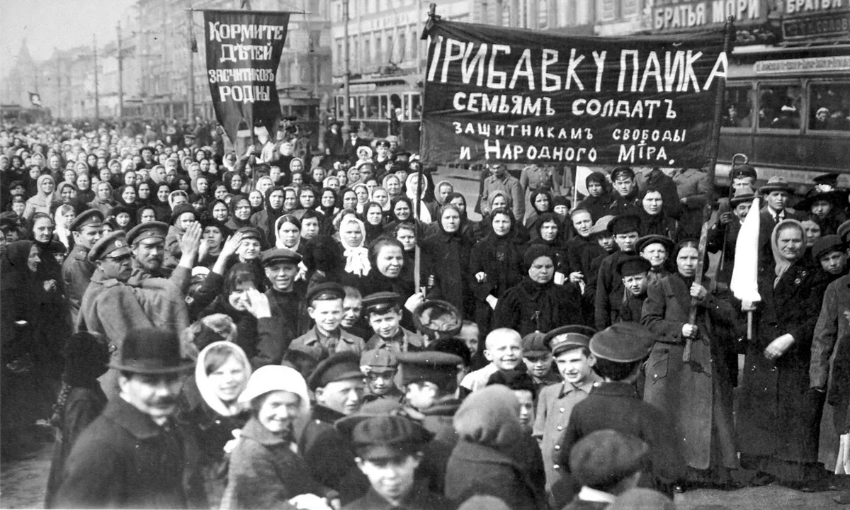 Black-and-white photo of a dense crowd of women in headscarves and men in caps under large banners with Cyrillic slogans on a city street with a tram.