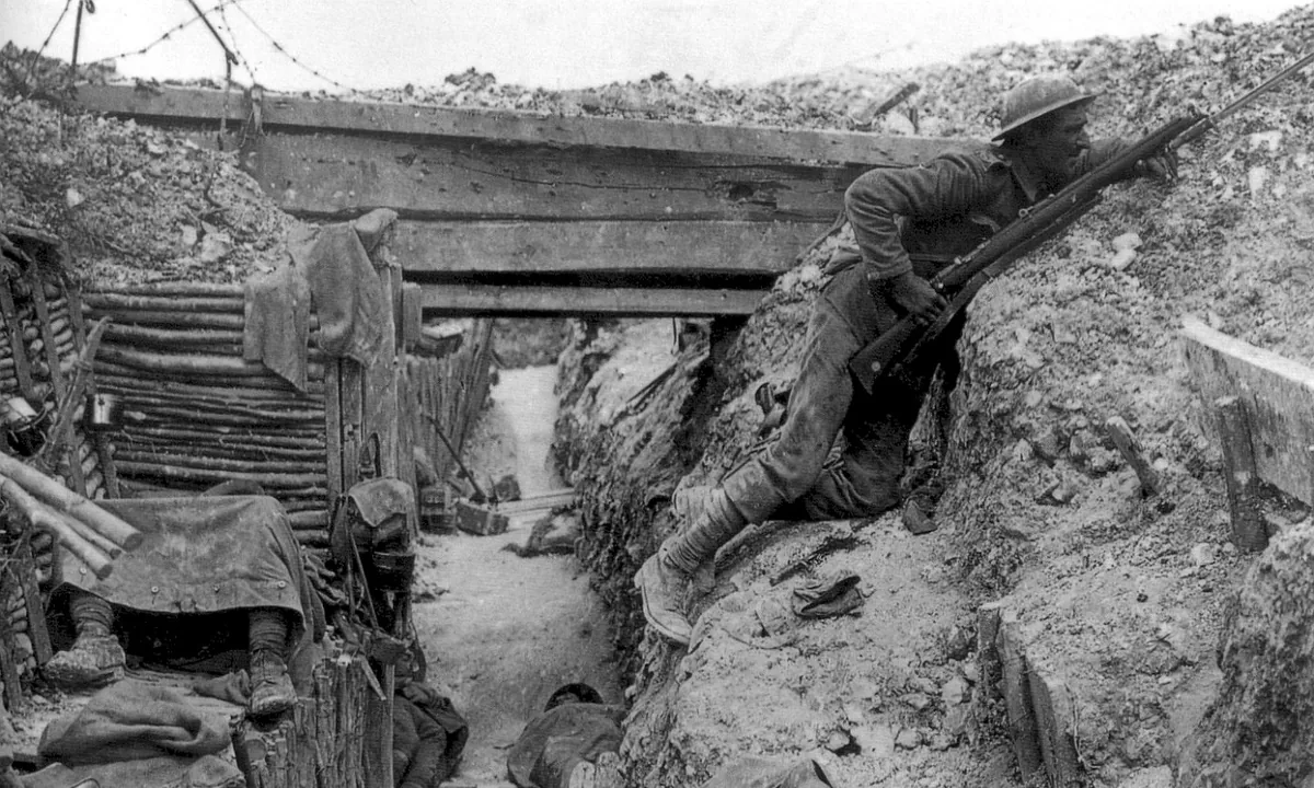 Black-and-white photo of a timber-lined trench: one soldier with a rifle watches over the parapet while others sleep under ground sheets.